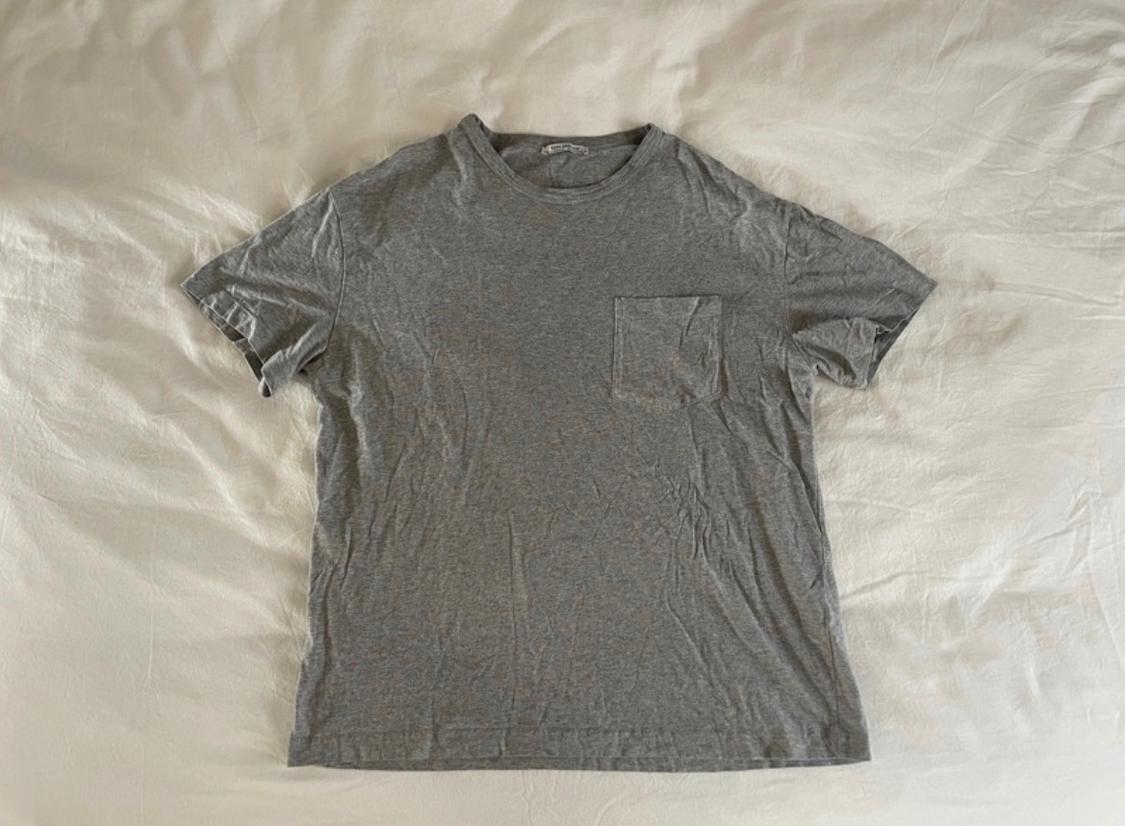 Basic grey short-sleeved T-shirt with a chest pocket, laid on a crumpled white sheet. Casual style.