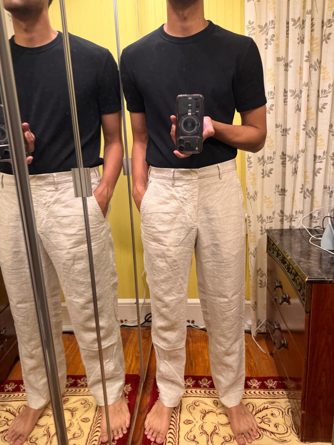 Man taking a selfie in the mirror. He is wearing a black t-shirt and beige linen pants. Relaxed atmosphere in a room with a rug.