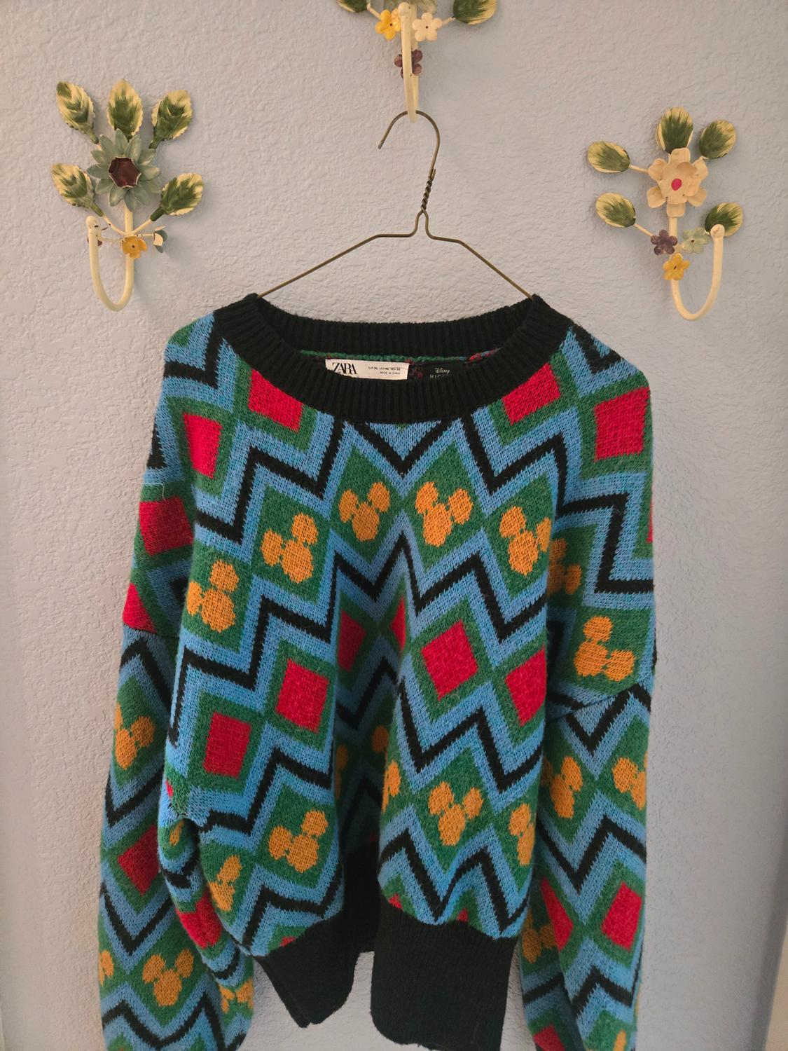 Colourful jumper with geometric patterns in blue, red, green, and yellow, hanging on a hanger against a light wall.