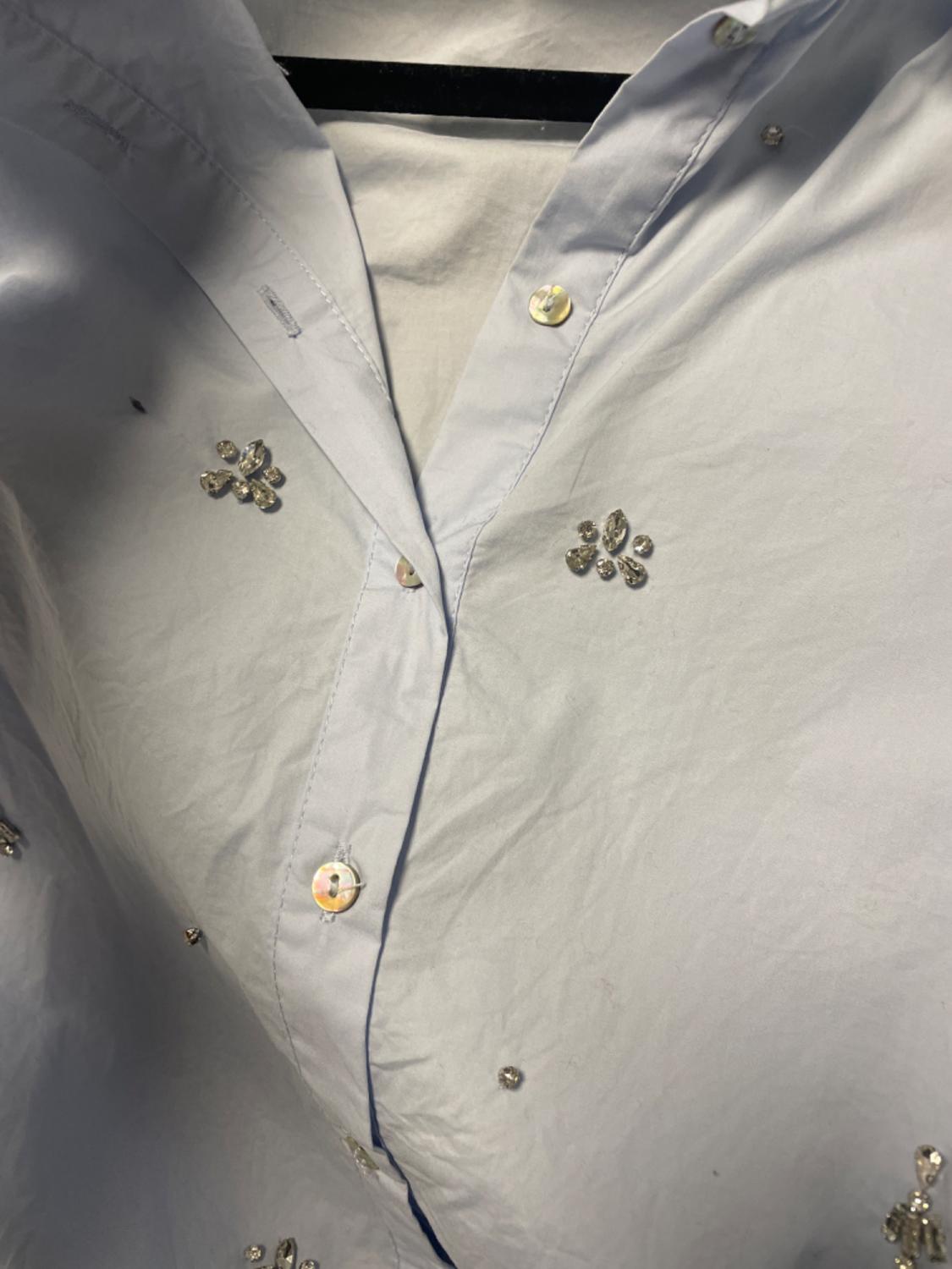 White shirt with shiny buttons and small floral rhinestone details, hanging on a black hanger.