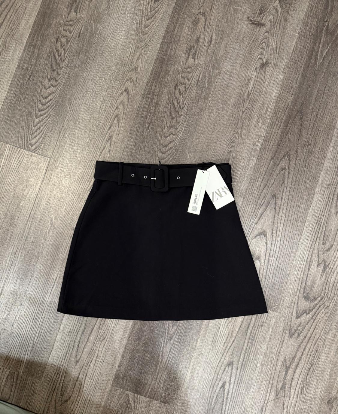 Short black skirt with a belt and store tag on a gray wooden floor.