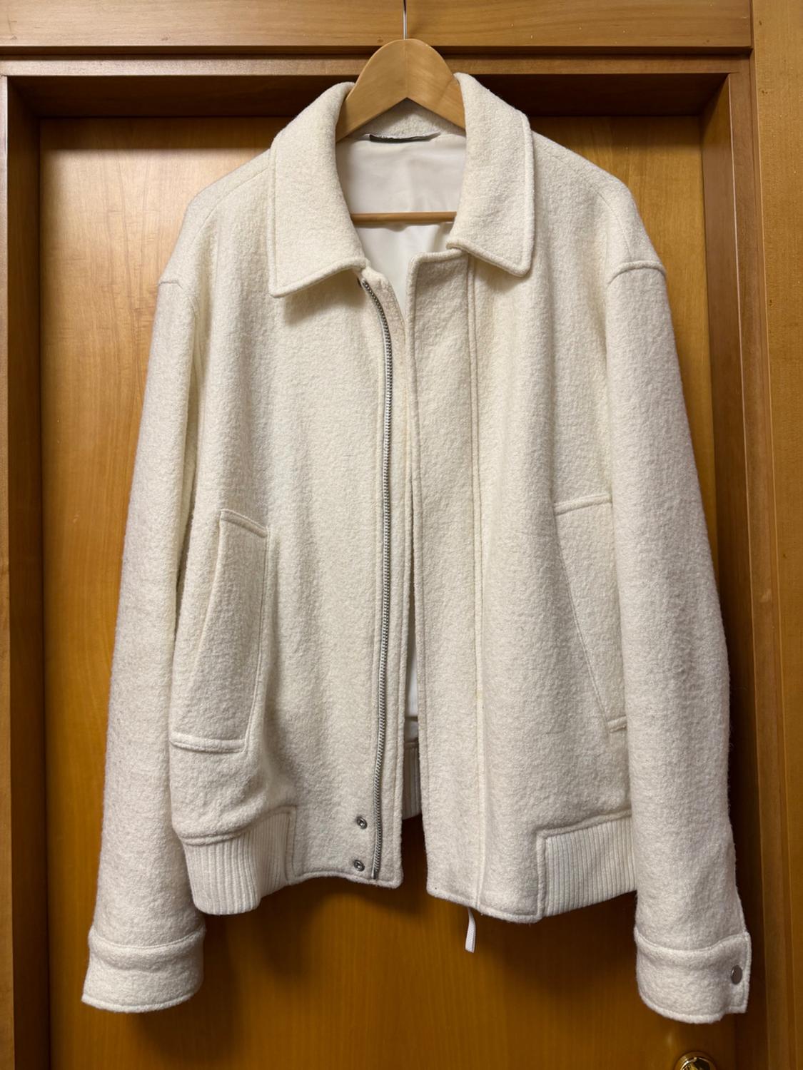 White wool jacket hanging on a wooden hanger, with a front zip and pockets, over a light wooden door.