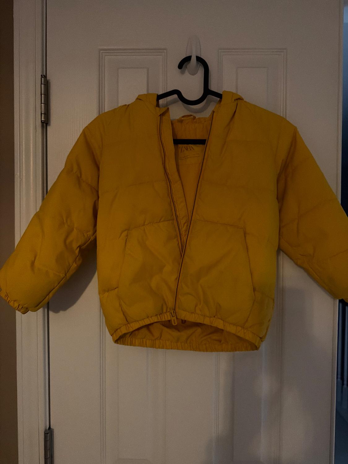 Yellow quilted jacket hanging on a white door. The jacket has a hood and a front zipper. Indoor setting.