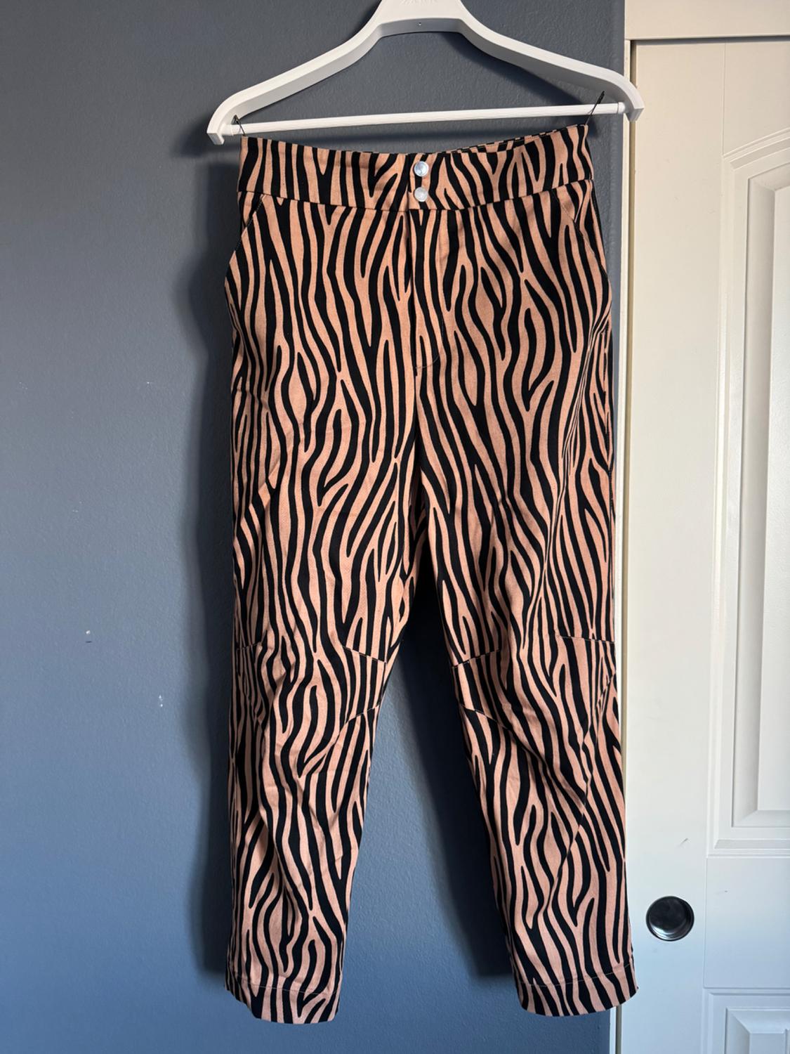 High-waisted pants with a black striped print on a beige background, hanging on a white hanger in front of a gray wall.
