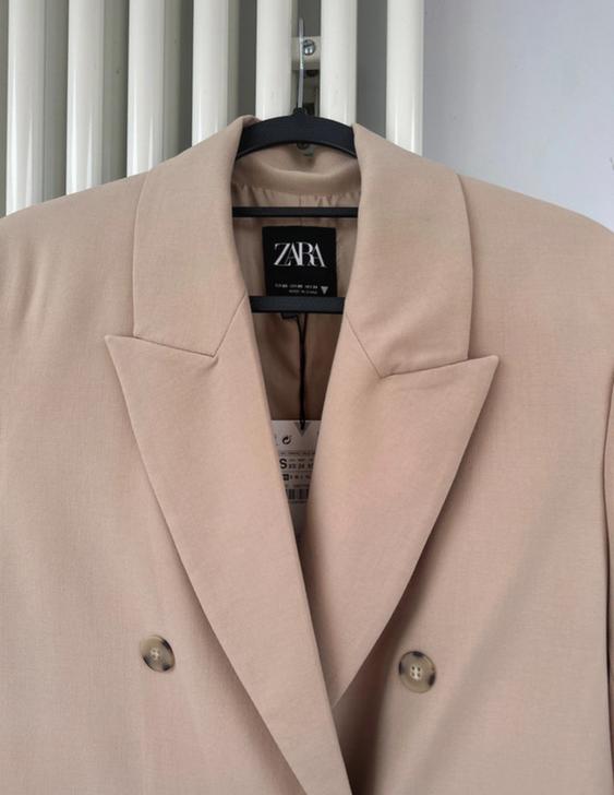 Double-breasted beige jacket hanging on a hanger, with a visible Zara label. White radiator in the background.