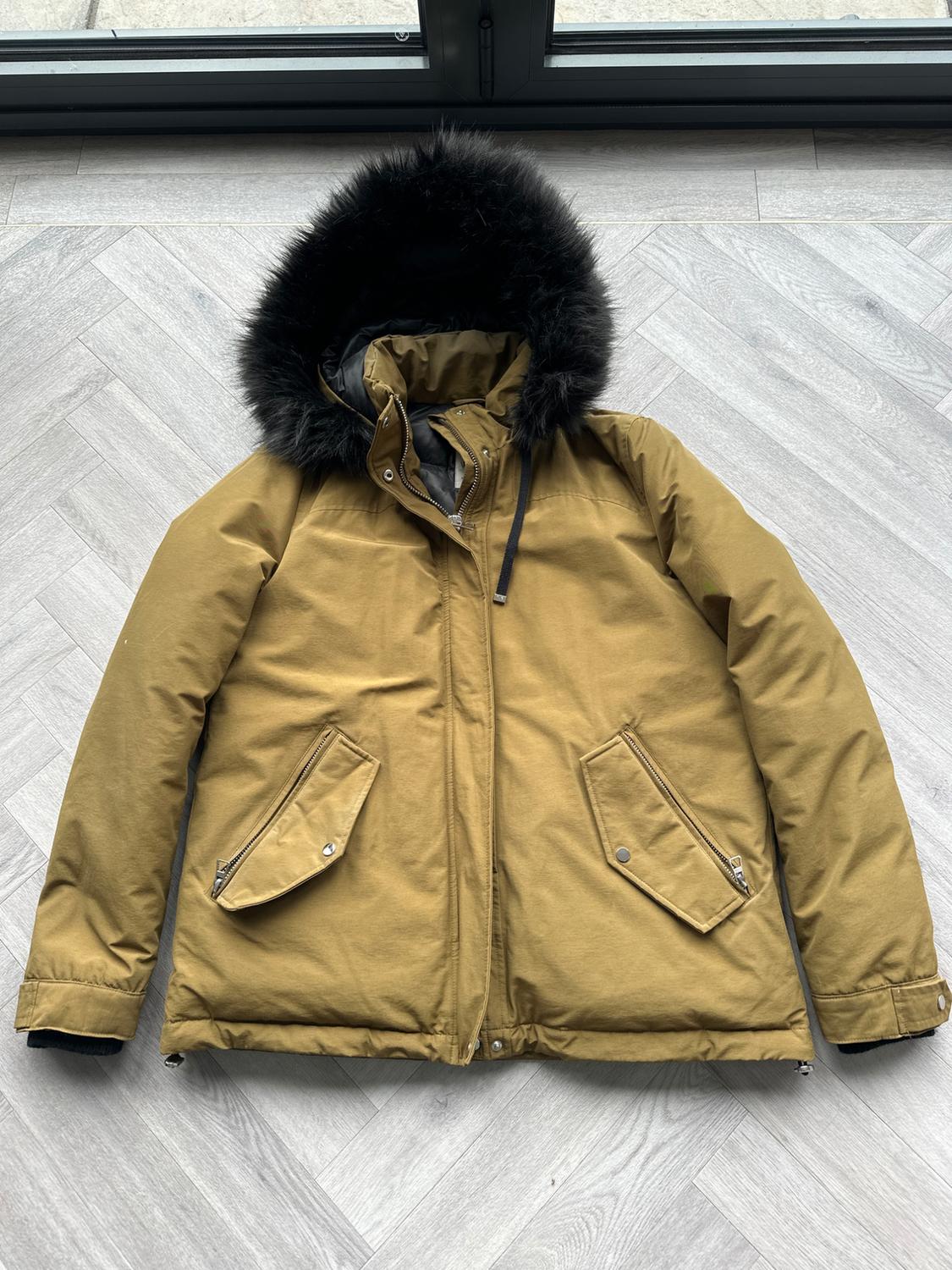 Brown jacket with a black fur-trimmed hood, flap pockets and zip fastening, on a light wood floor.