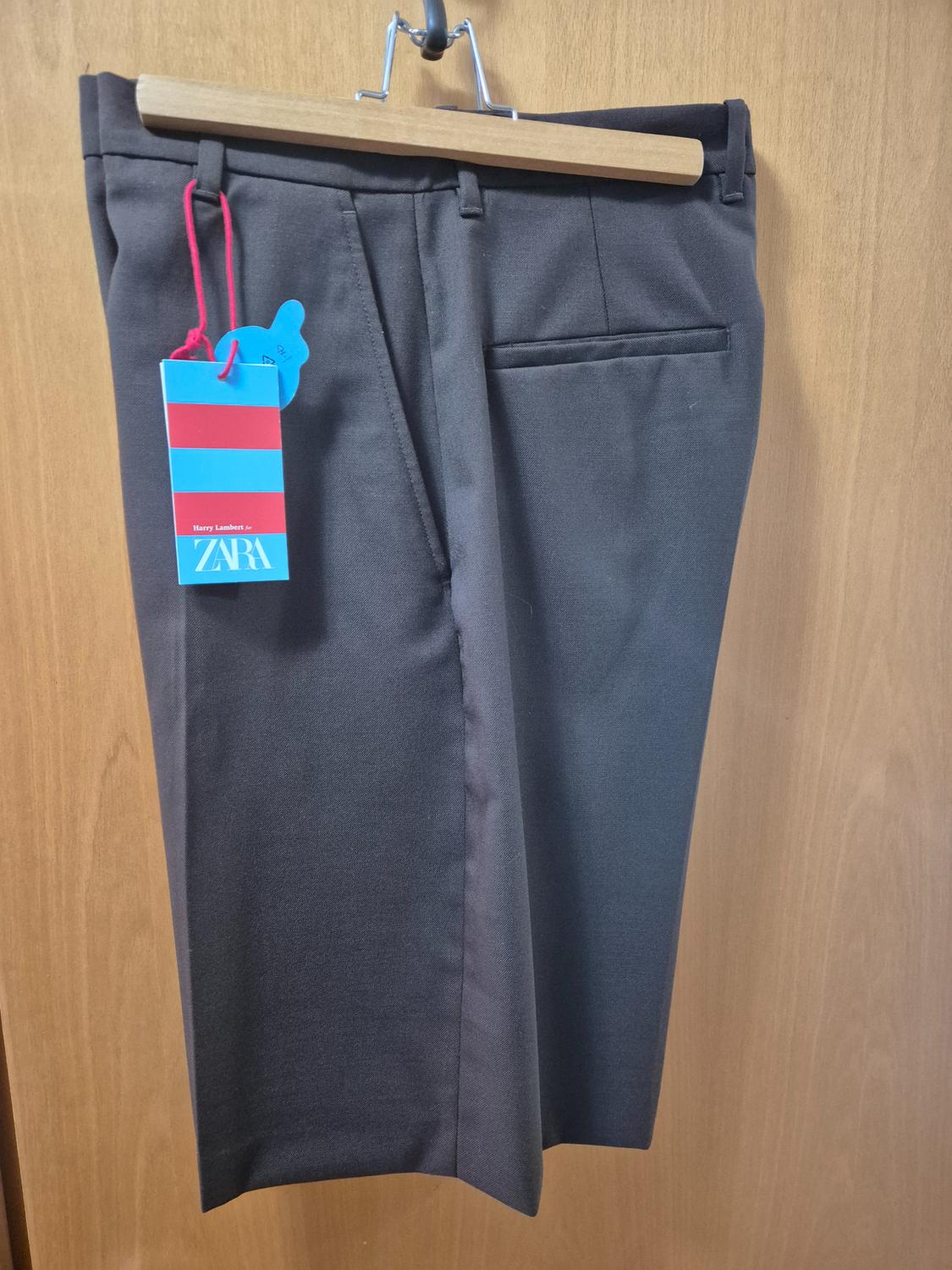 Grey trousers hanging on a wooden hanger with a blue and red label that says "ZARA" against a light wood background.