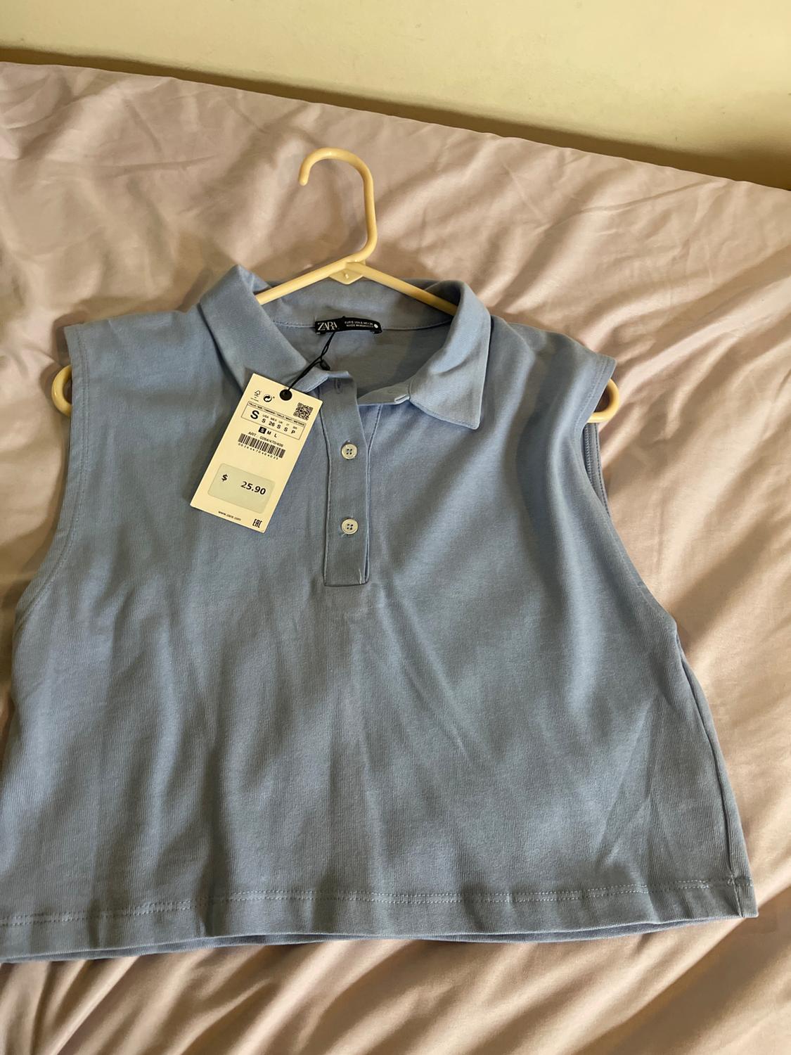 Light blue sleeveless top with a polo collar and buttons, hanging on a hanger over a bed. Visible tag with a price of $25.90.