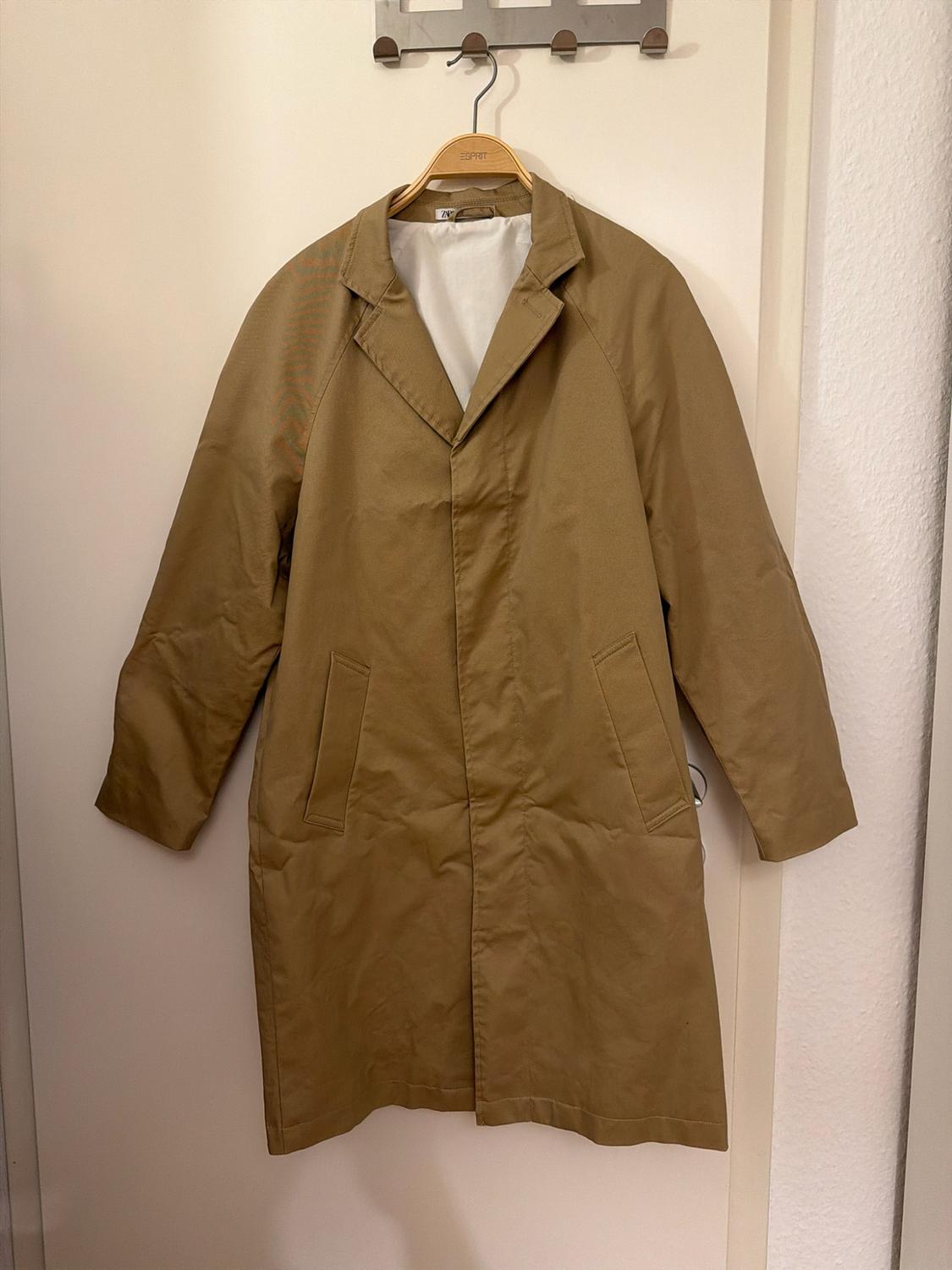 Long camel-coloured coat hanging on a wooden hanger bearing the text ESPRIT, on a white door beneath a coat rack.