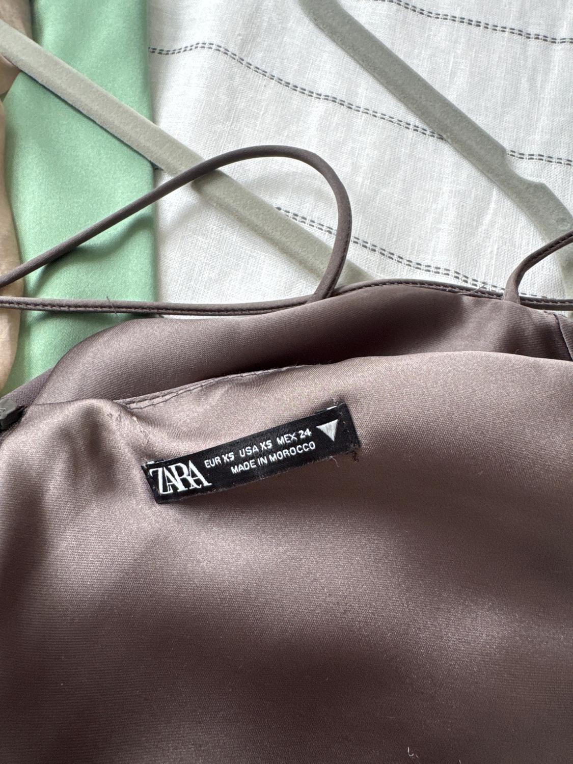 Zara clothing label on brown fabric, reads: "EUR XS USA XS MEX 24 MADE IN MOROCCO". Background with green, beige, and white striped fabrics.