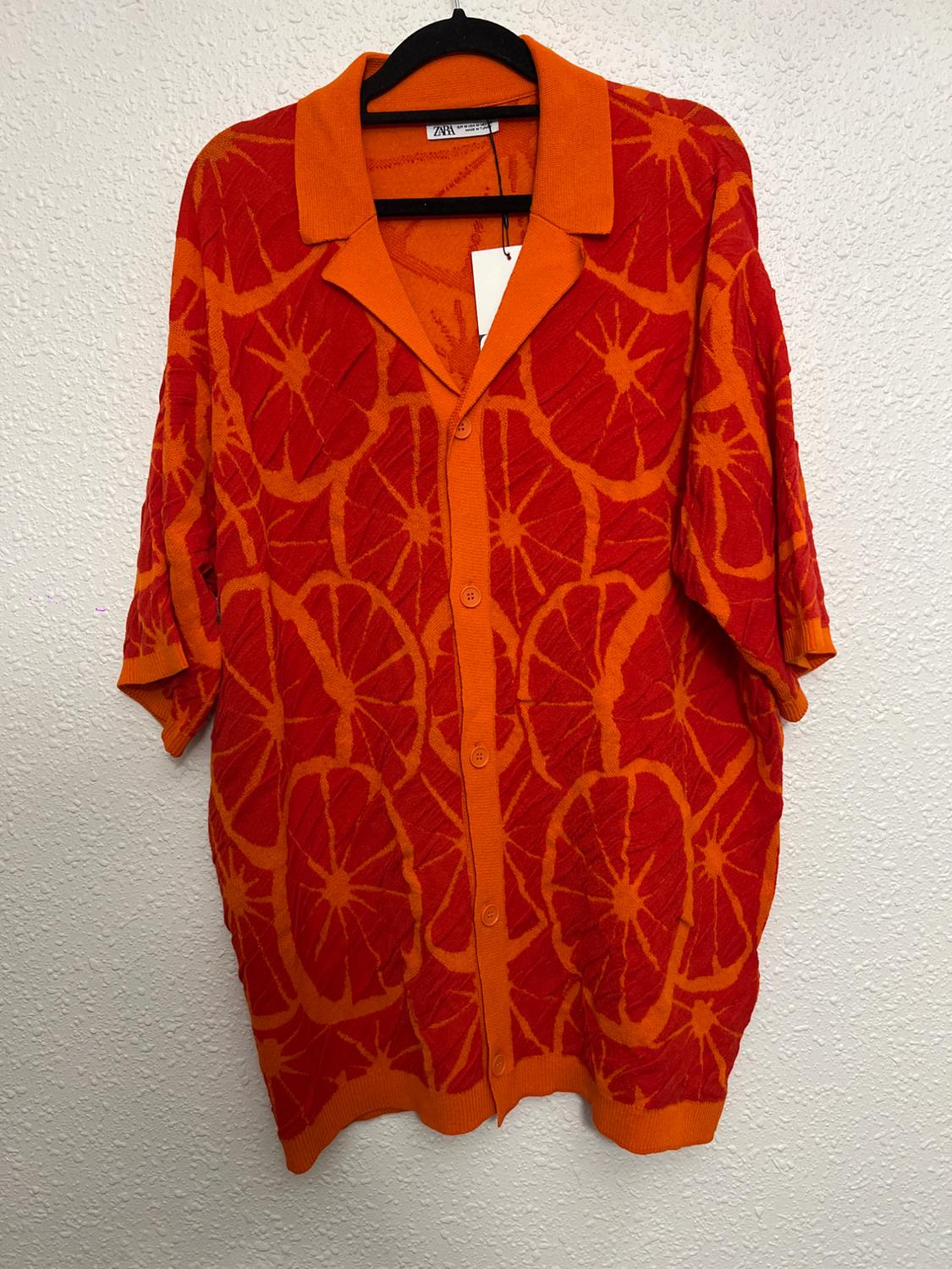 Orange and red cardigan with an orange slice print, hanging on a black hanger against a white wall.
