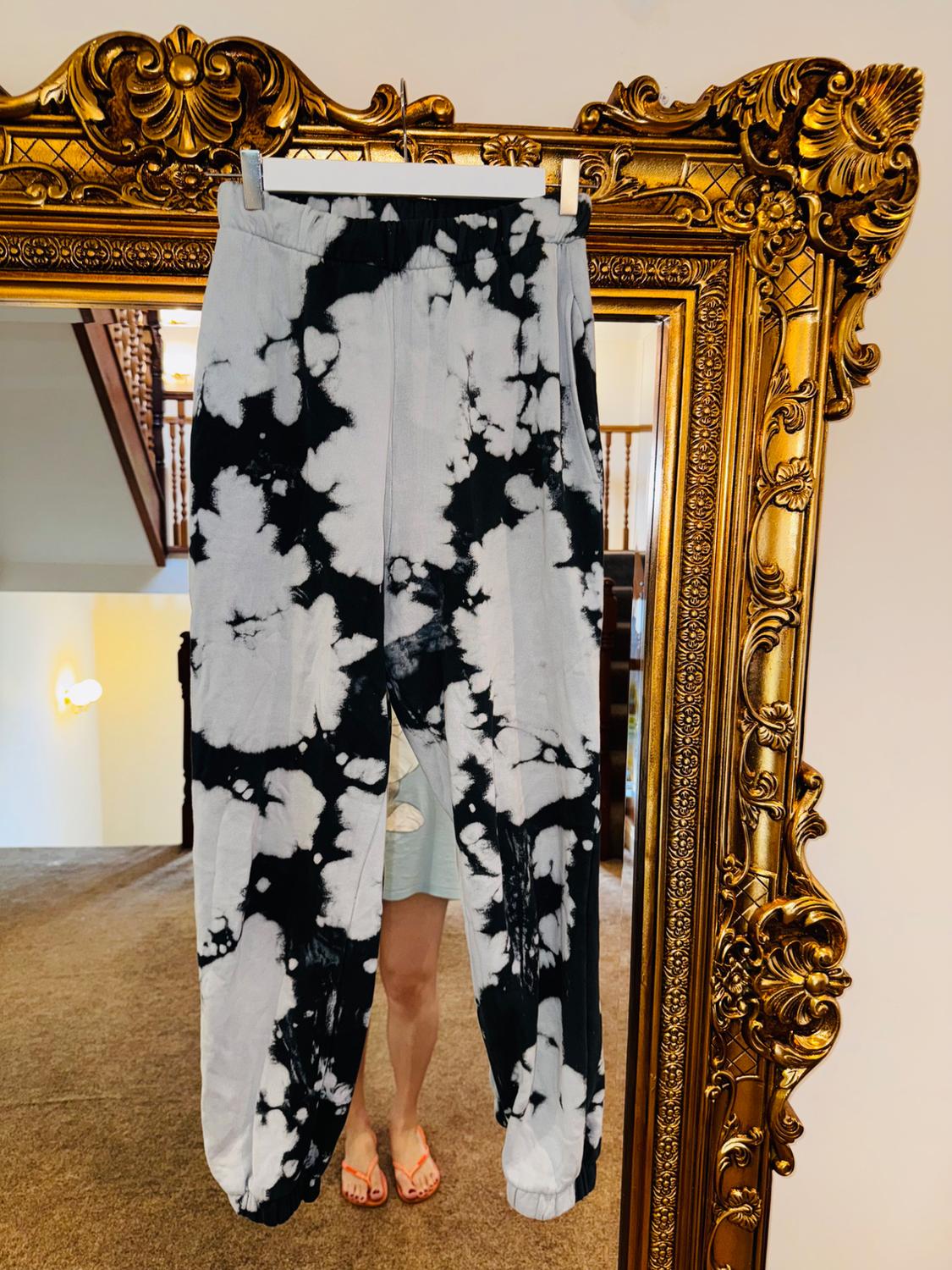 Black and white tie-dye design trousers hanging on an ornate gold mirror. Person wearing sandals in the background.