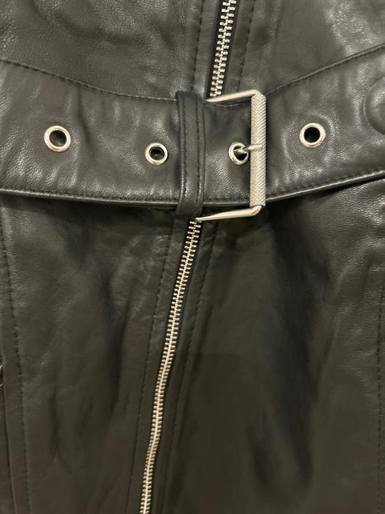 Detail of a black jacket with a metallic zip and a belt with a silver buckle and eyelets. Soft textured fabric.