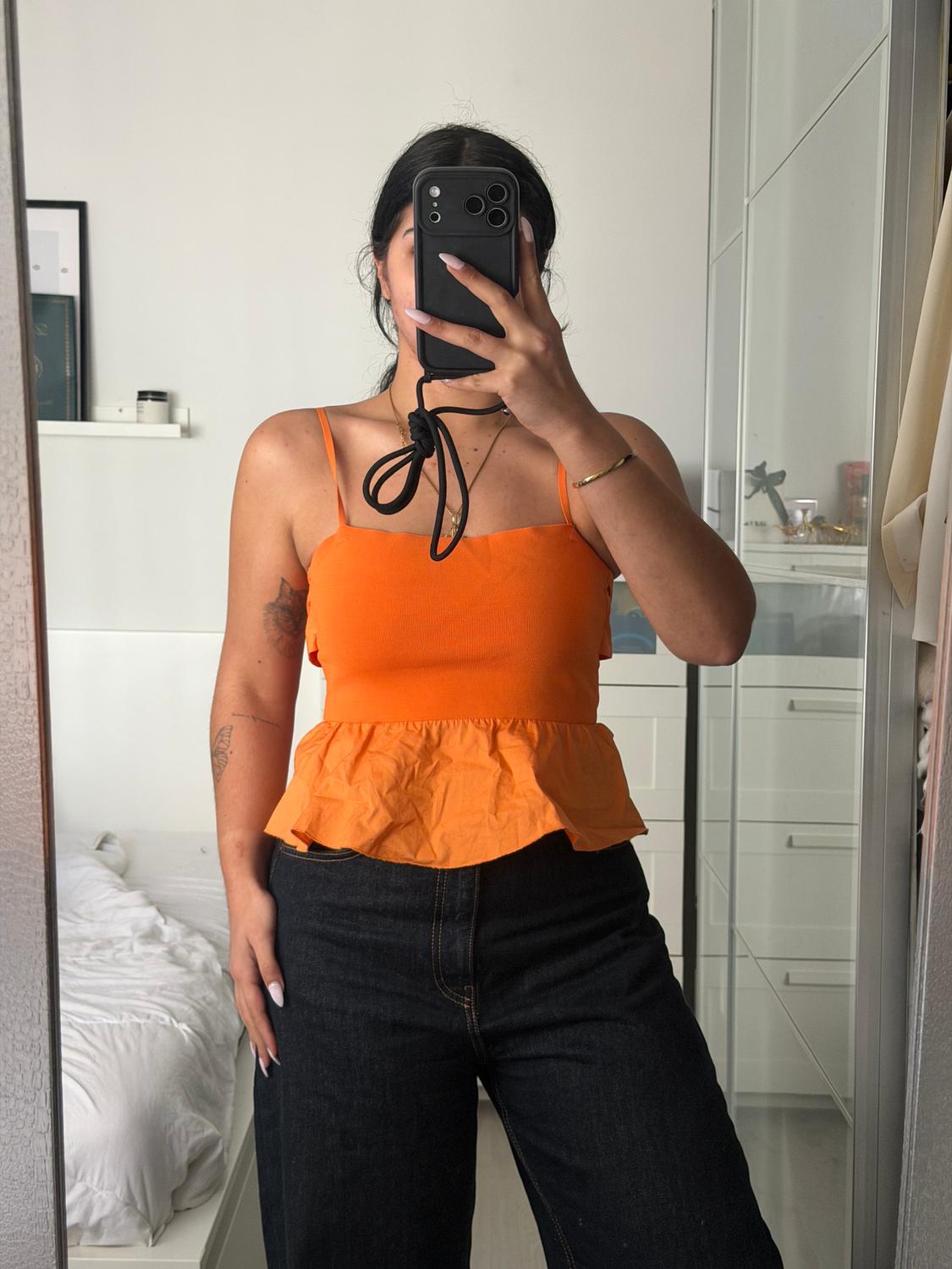 Woman in an orange spaghetti strap top and dark jeans, taking a photo in front of a mirror in a modern bedroom.