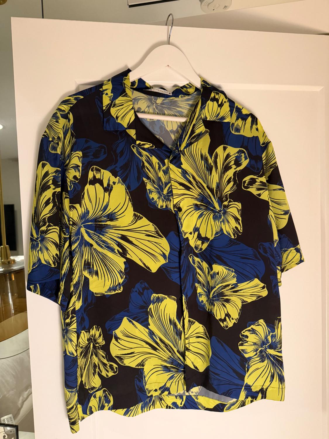 Short-sleeved shirt with a large yellow and blue floral print on a dark background, hanging on a white hanger on a door.