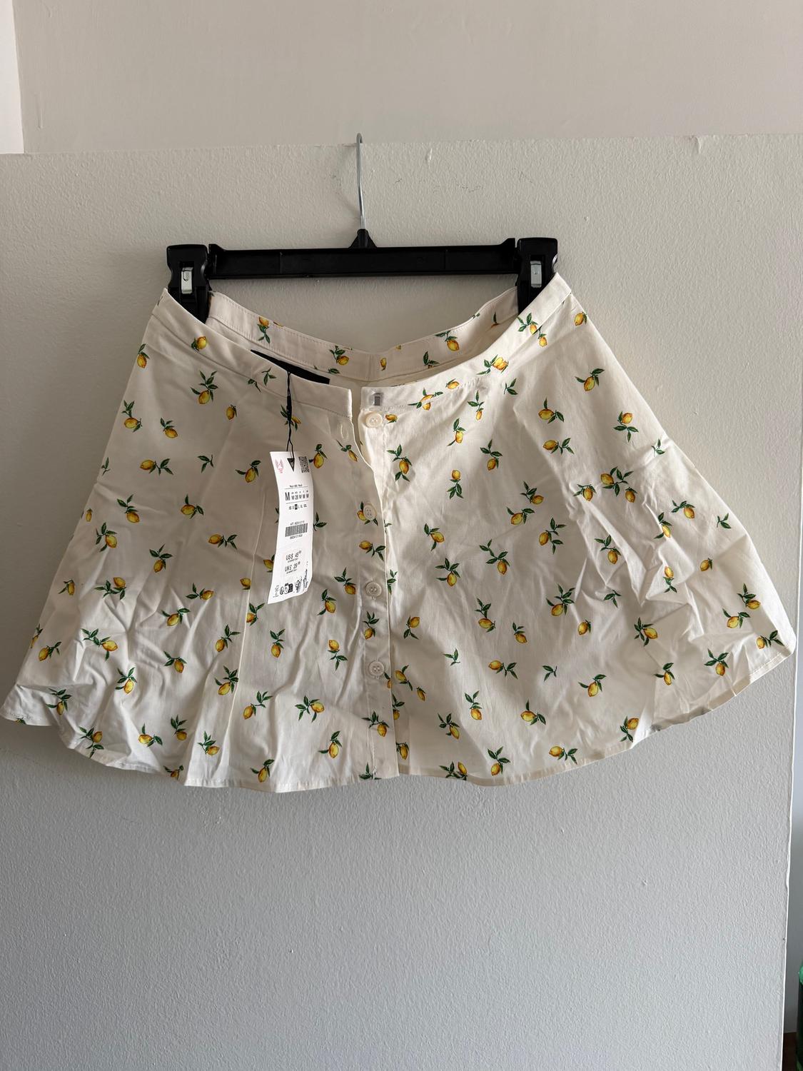 White skirt with a yellow and green lemon print, hanging on a black hanger. Visible label with text in English.