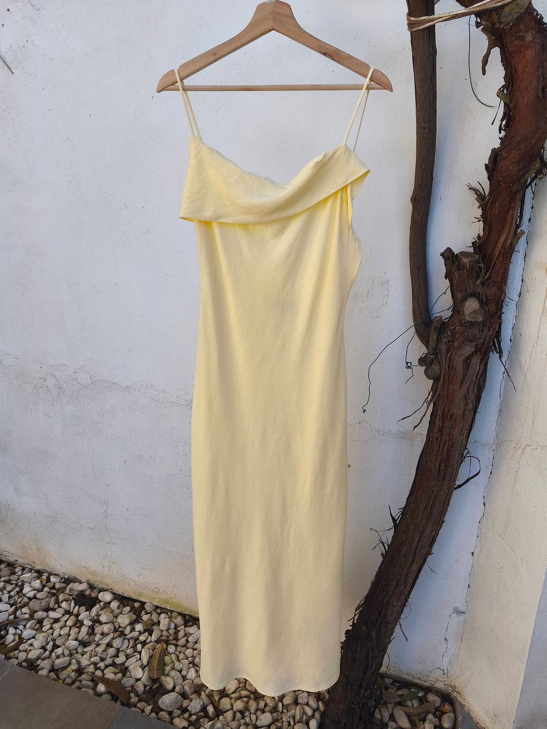 Long yellow dress with thin straps hanging on a wooden hanger, next to a white wall and a dry branch.