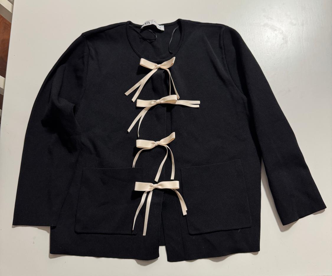 Black jacket on a white surface. Long-sleeved design, round neck, two pockets and front fastening with four cream-coloured ties.