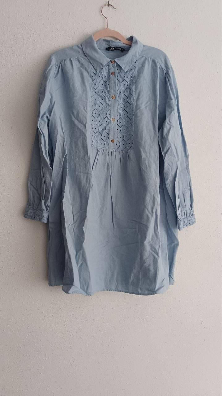 Light blue shirt with embroidery on the chest and wooden buttons, hanging on a hanger against a white wall.