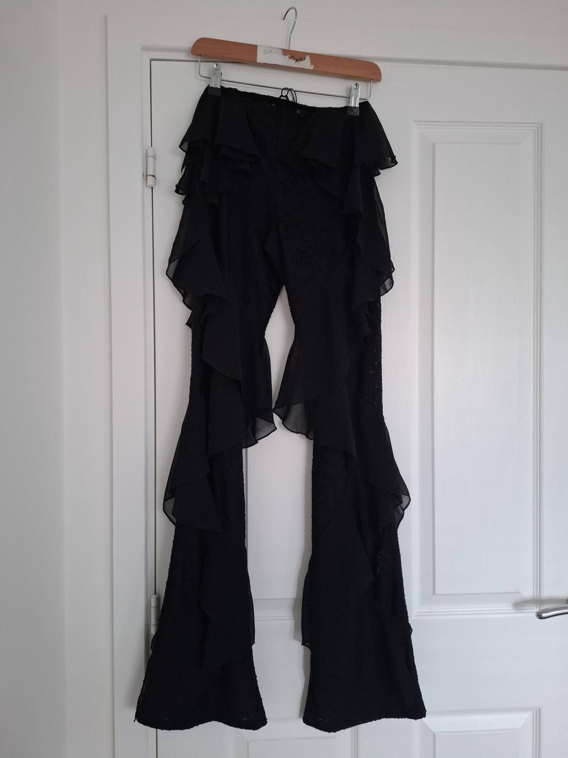 Black trousers with side ruffles hanging from a wooden hanger on a white door.