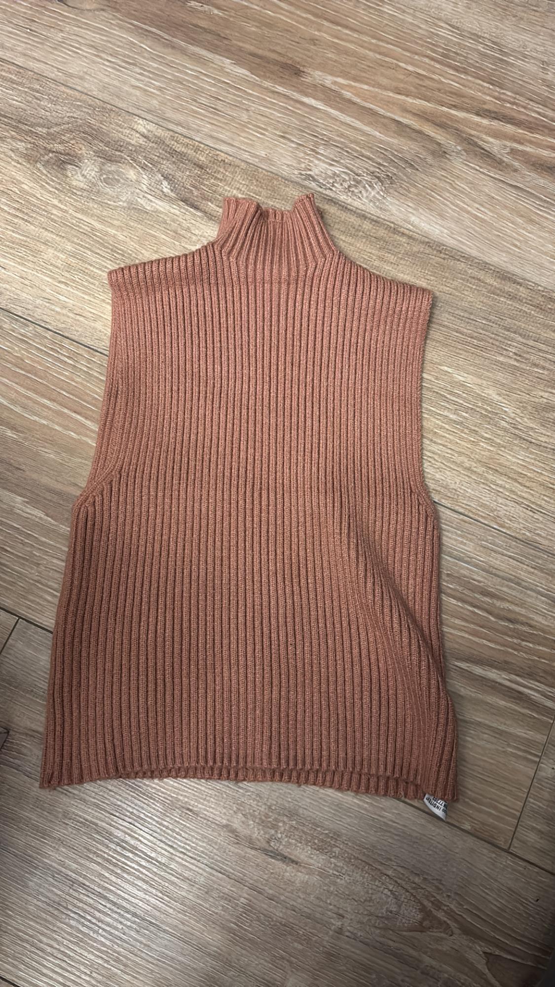 Terracotta ribbed knit sleeveless vest on a light wooden floor. High neck and simple design.