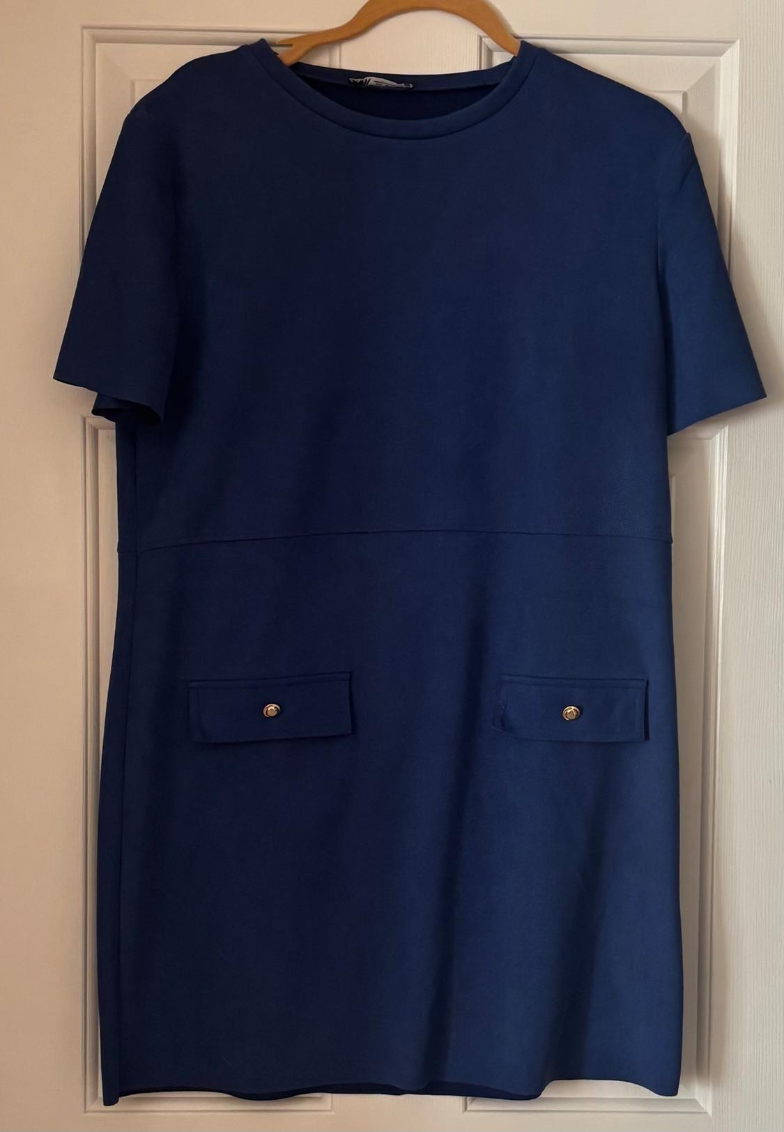 Blue short-sleeved dress hanging on a wooden hanger. It has two front pockets with gold buttons.