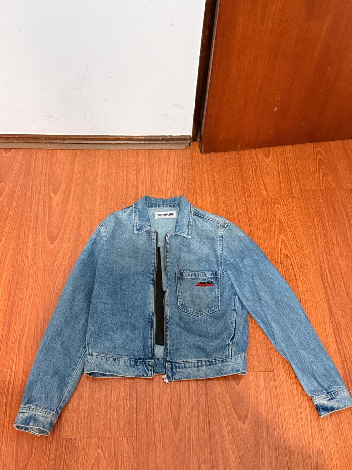 Light blue denim jacket with zip, chest pocket and "RHUGI" label. Wooden floor and white wall background.