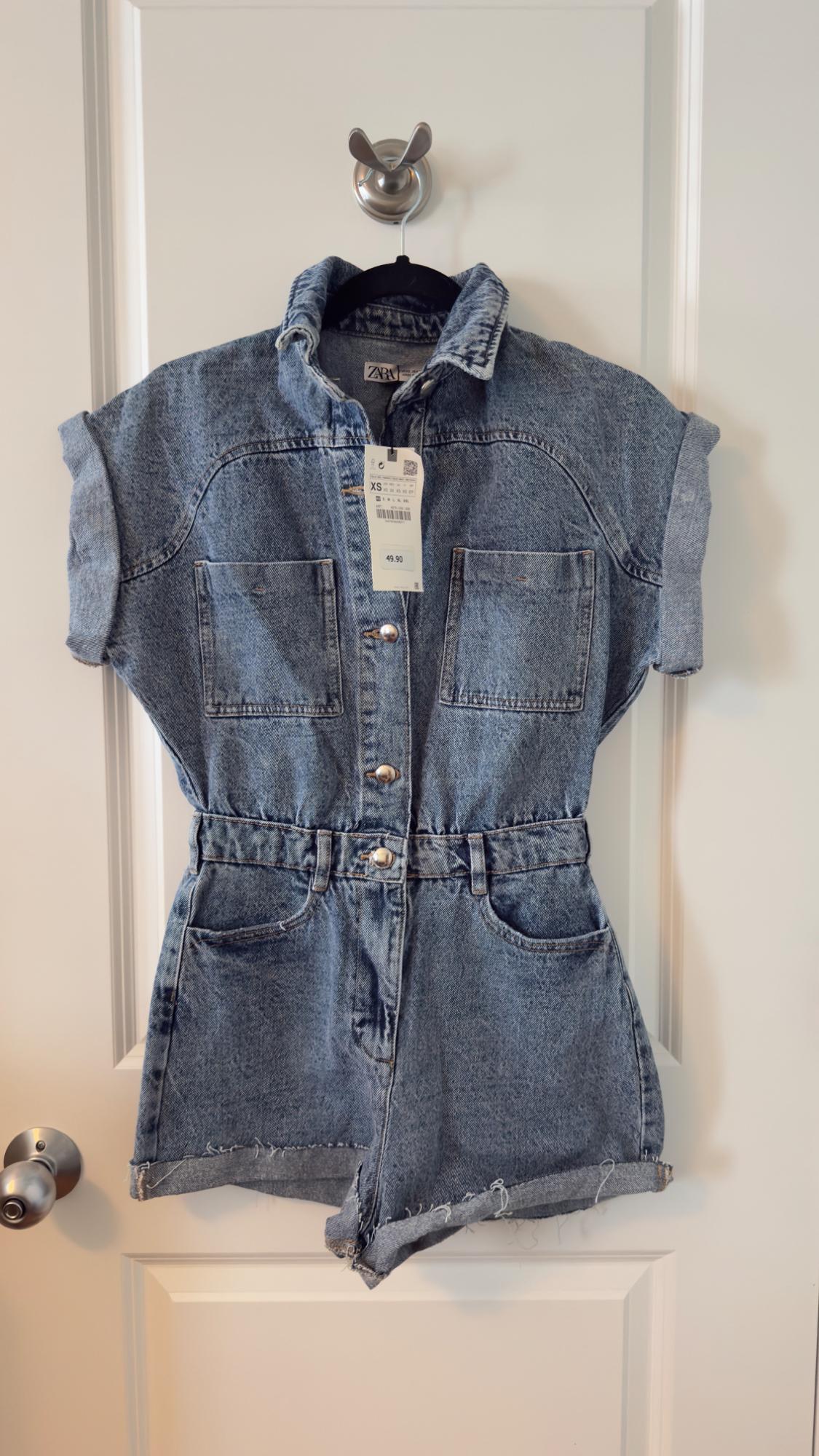 Blue denim jumpsuit with short sleeves and front pockets, hanging on a white door. Visible tag with price of 49.90.