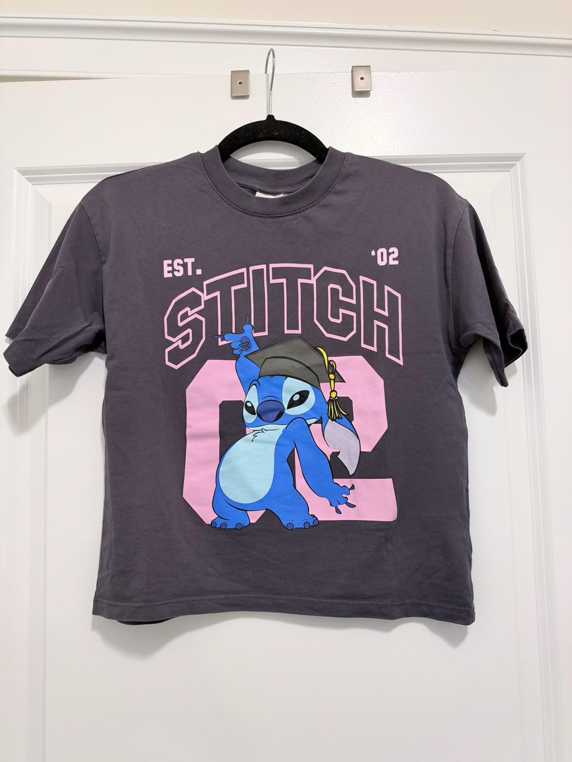 Dark gray t-shirt hanging on a white door. Stitch print with a graduation cap and the text "EST. STITCH '02" over a pink "02".