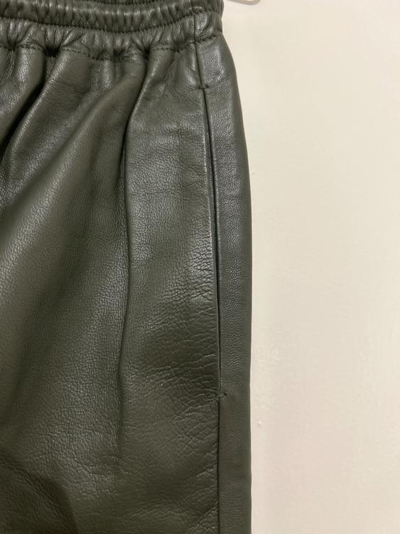 Detail of dark green leather trousers with elasticated waist and visible stitching, on a white background.