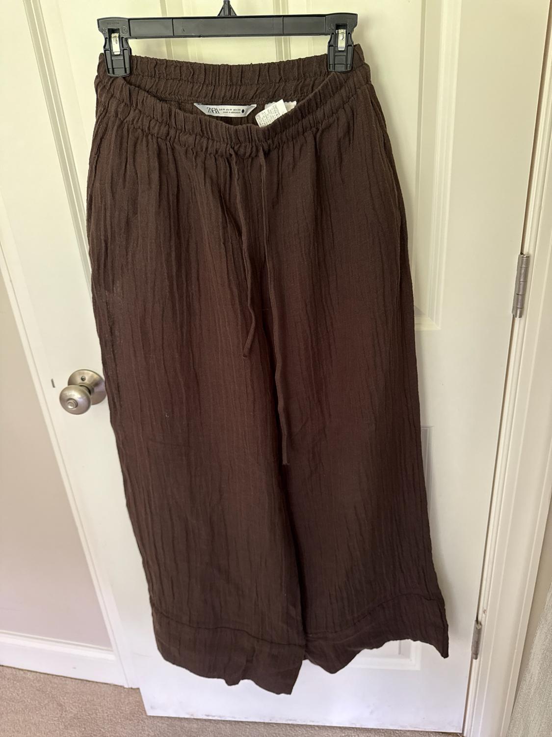 Brown wide-leg pants with texture, an elastic waistband, and drawstring, hanging on a white door. ZARA label visible.