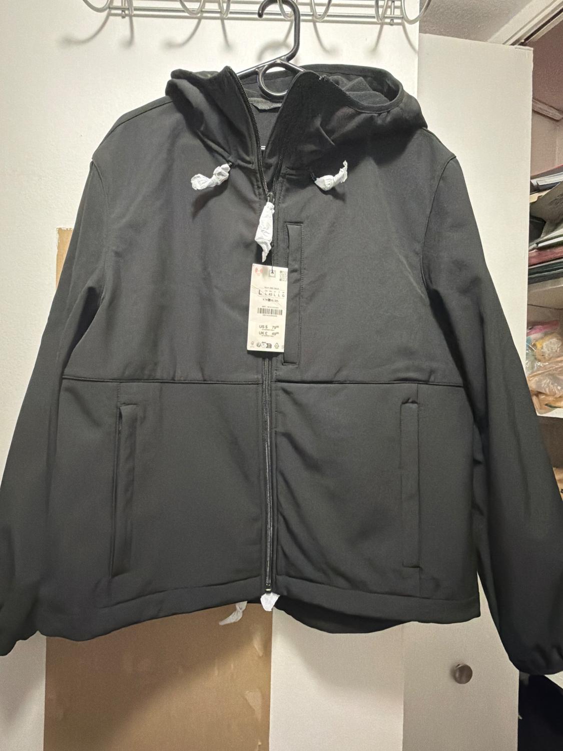 Black jacket with a hood hanging on a hanger. Design with a zipper closure, front pockets, drawstrings, and a price tag.