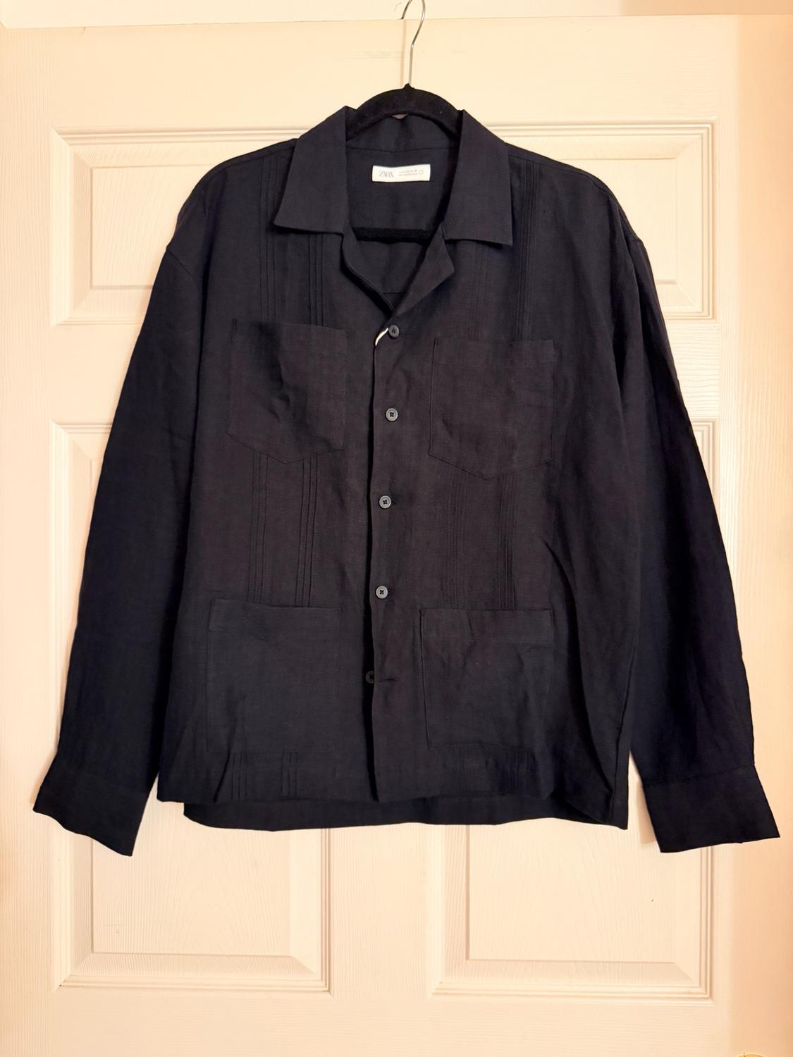 Black long-sleeved shirt with pleats and four pockets, hanging on a white door. Label with the text ZARA on the collar.