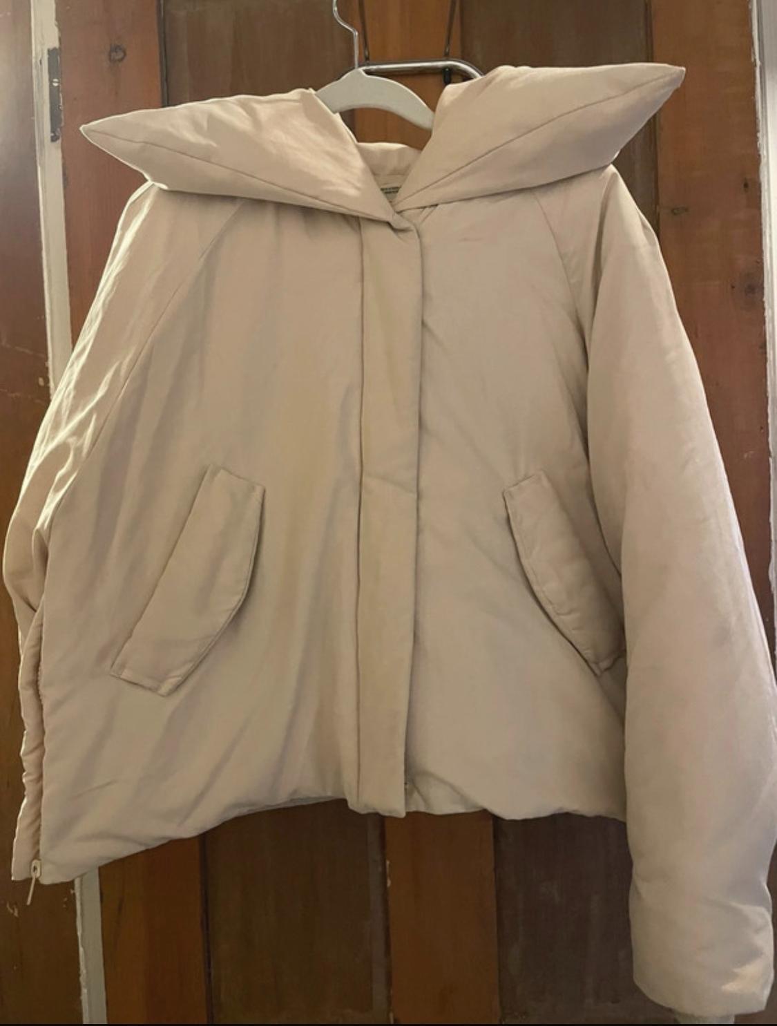 Beige padded coat with a wide collar and pockets, hanging on a hanger over a wooden door.