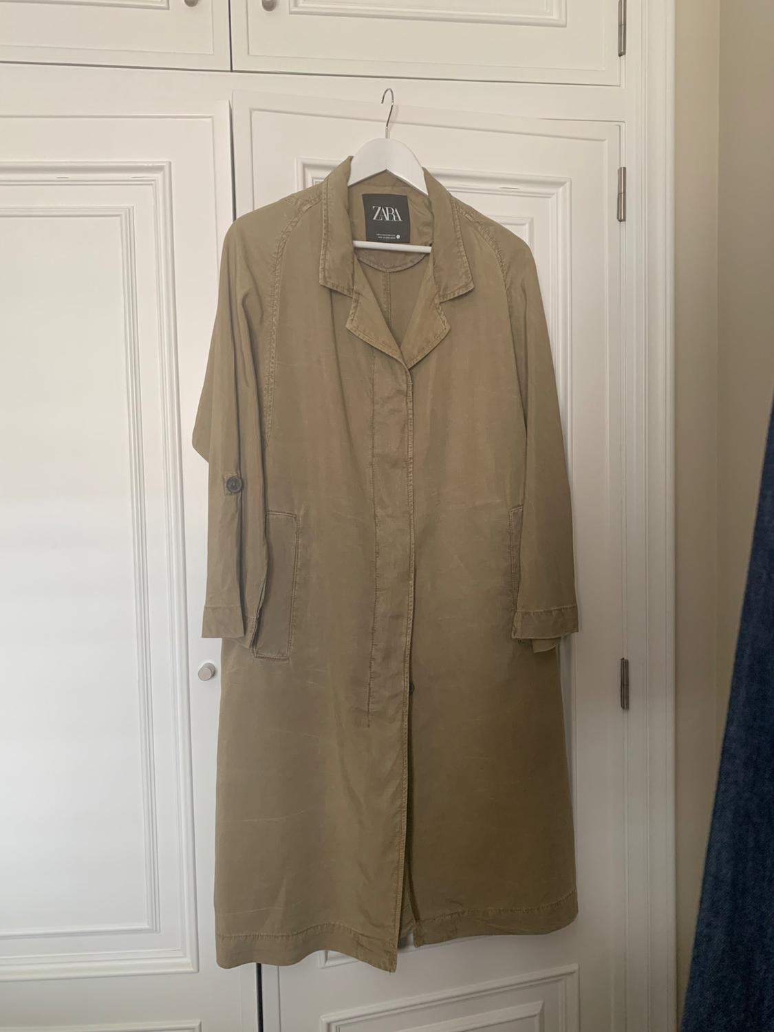 Long beige coat hanging on a white hanger in front of a white wardrobe. Visible label with the text "ZARA".
