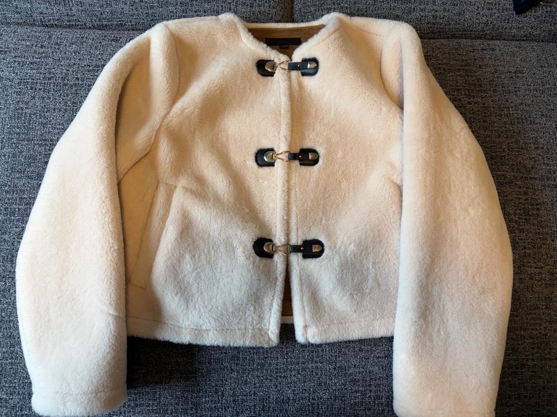 White wool jacket with black buckle fastenings, on a grey background. Long-sleeved and straight-cut design.