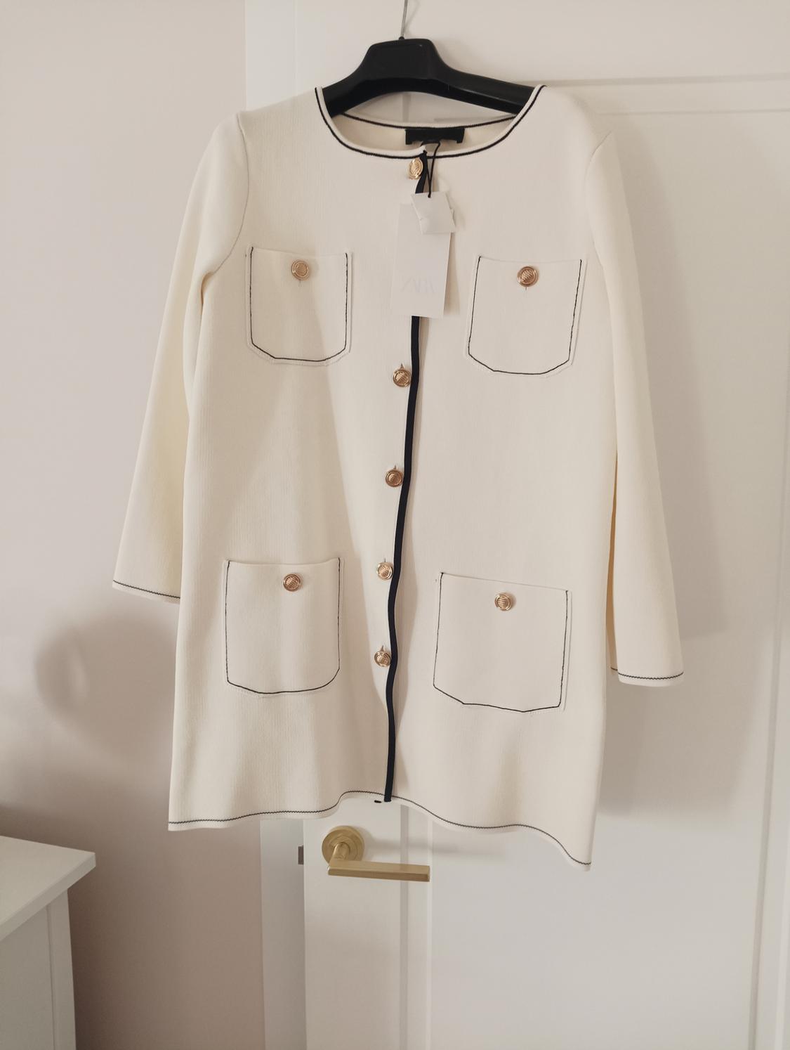 White coat with gold buttons and black trim, hanging on a black hanger over a white door. Label visible.