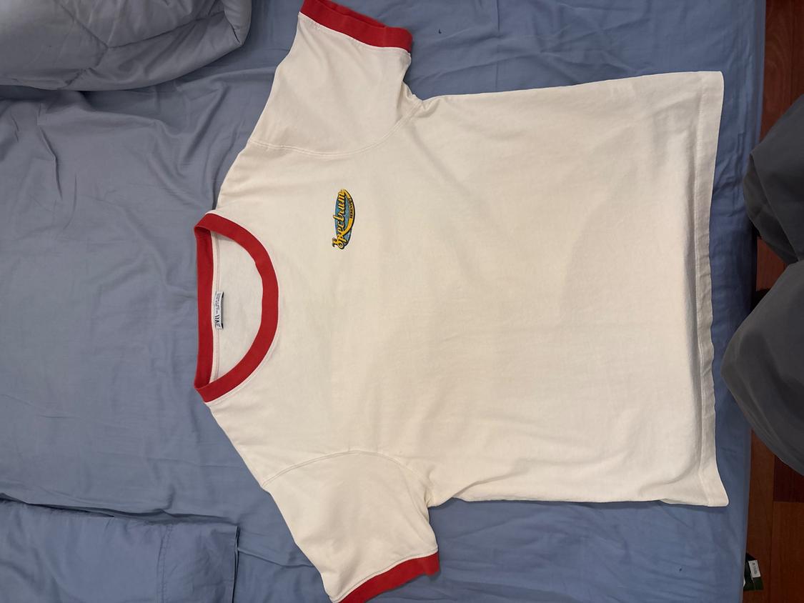 White T-shirt with red trim on the collar and sleeves, on a blue bed. Colourful logo on the chest.