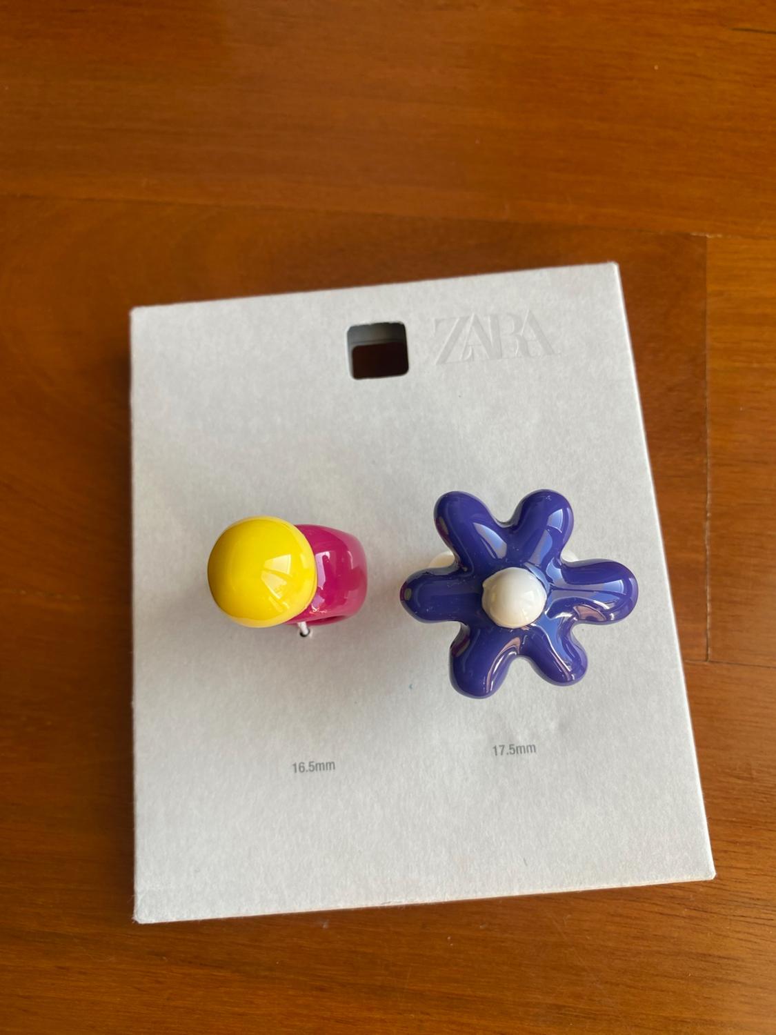 Two cardboard rings on wood. Text: ZARA, 16.5mm, 17.5mm. One ring is pink with a yellow sphere and the other is a purple flower.