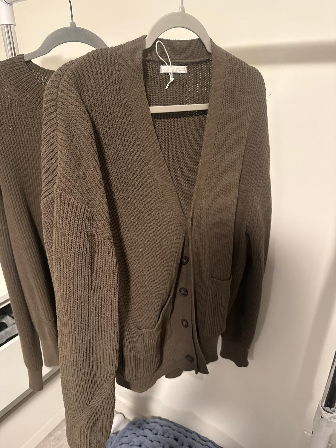 Brown knitted cardigan hanging on a grey hanger, with buttons and front pockets, against a light background.