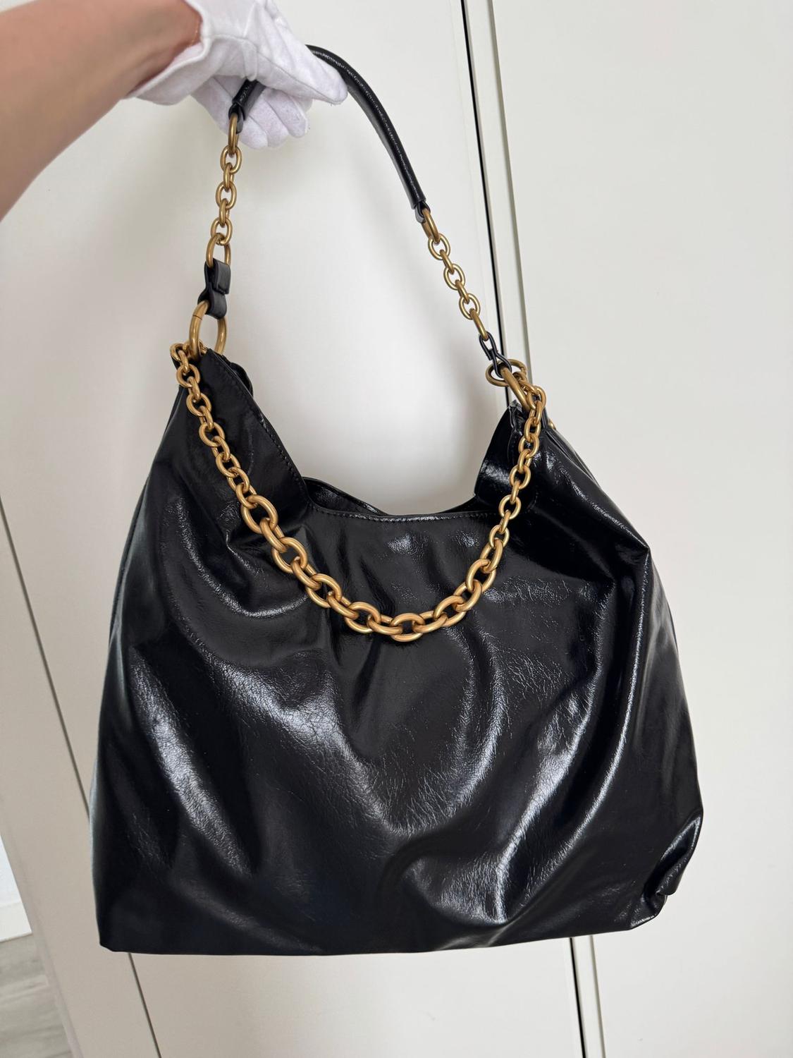 A shiny black handbag with a gold chain strap is held by a white gloved hand, with a white door in the background.