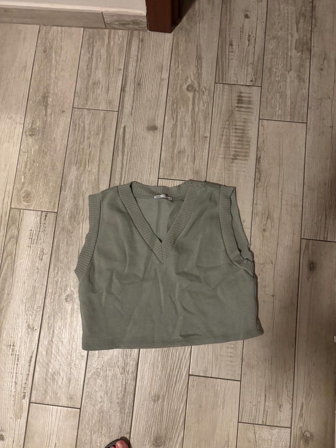 Light green knitted waistcoat with a V-neck, placed on a light wooden floor in a relaxed-style composition.