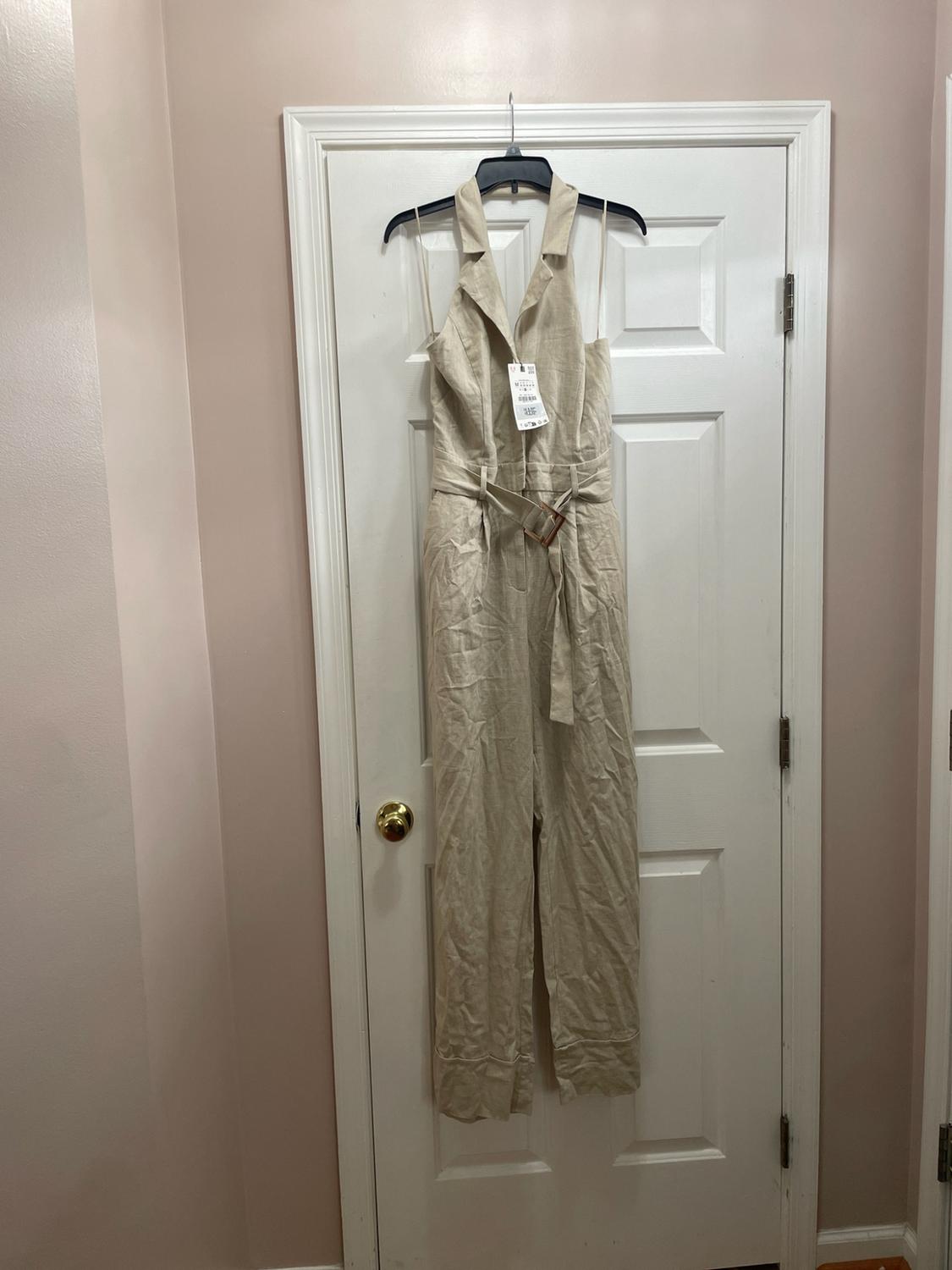 Long beige jumpsuit with a notched collar and belt, hanging on a hanger over a white door.