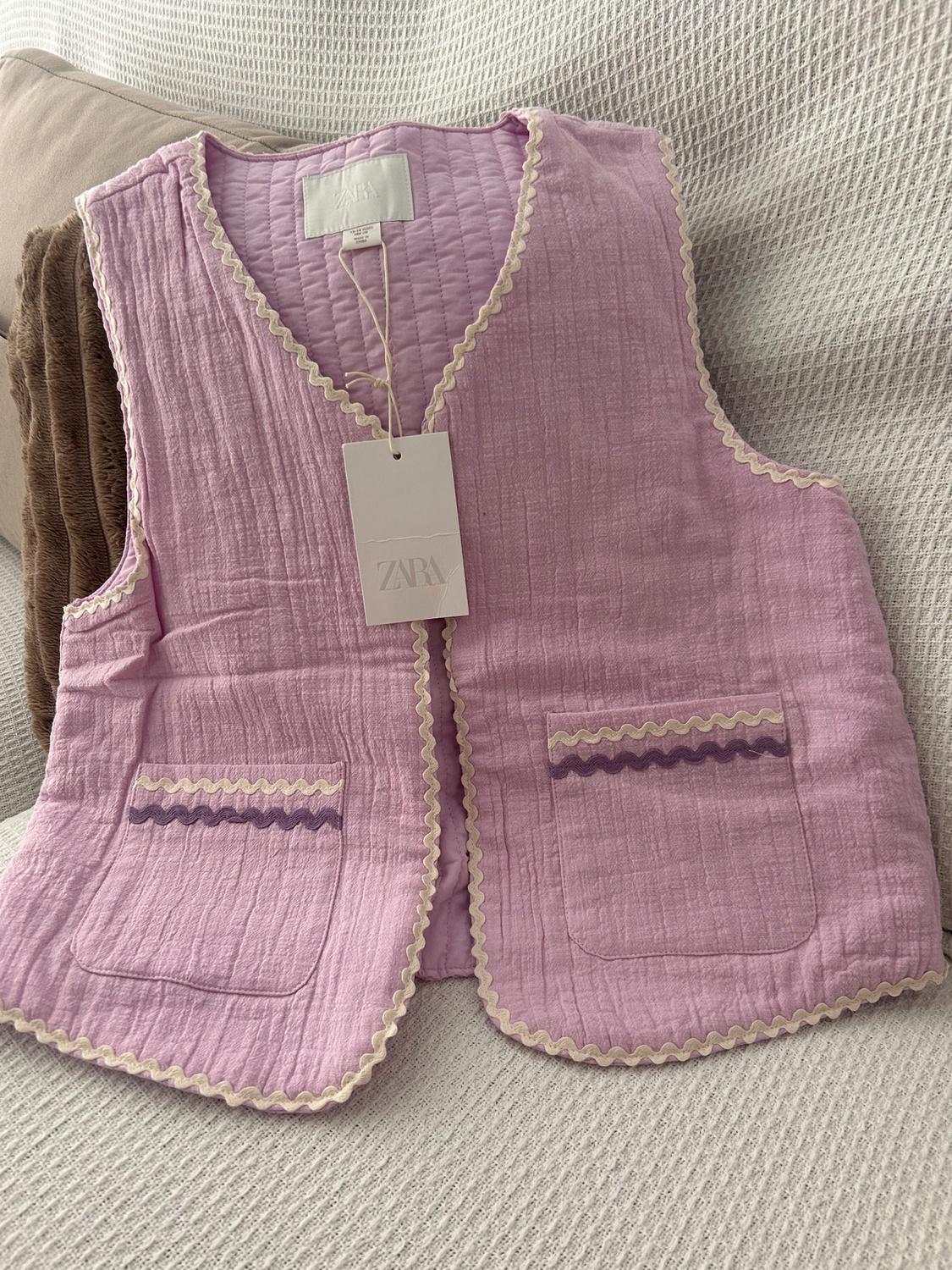 Lilac padded gilet with beige wavy trim and front pockets. Swing tag with the text "ZARA". Beige fabric background.