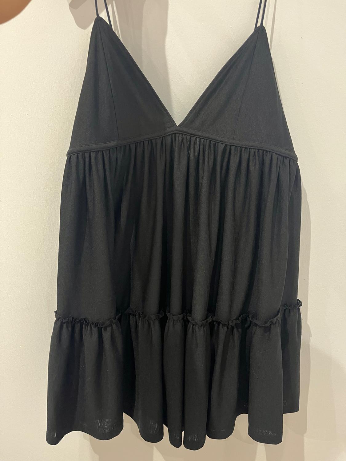 Black mini dress with spaghetti straps, V-neckline, gathered waist and frilled hem, hanging against a white wall.