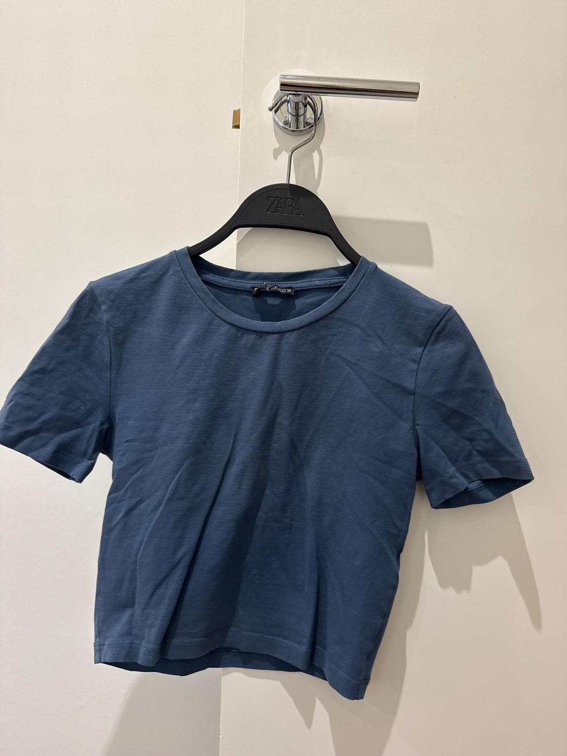Blue short-sleeved T-shirt hanging on a black hanger with the "Zara" logo, over a white door with a metal handle.