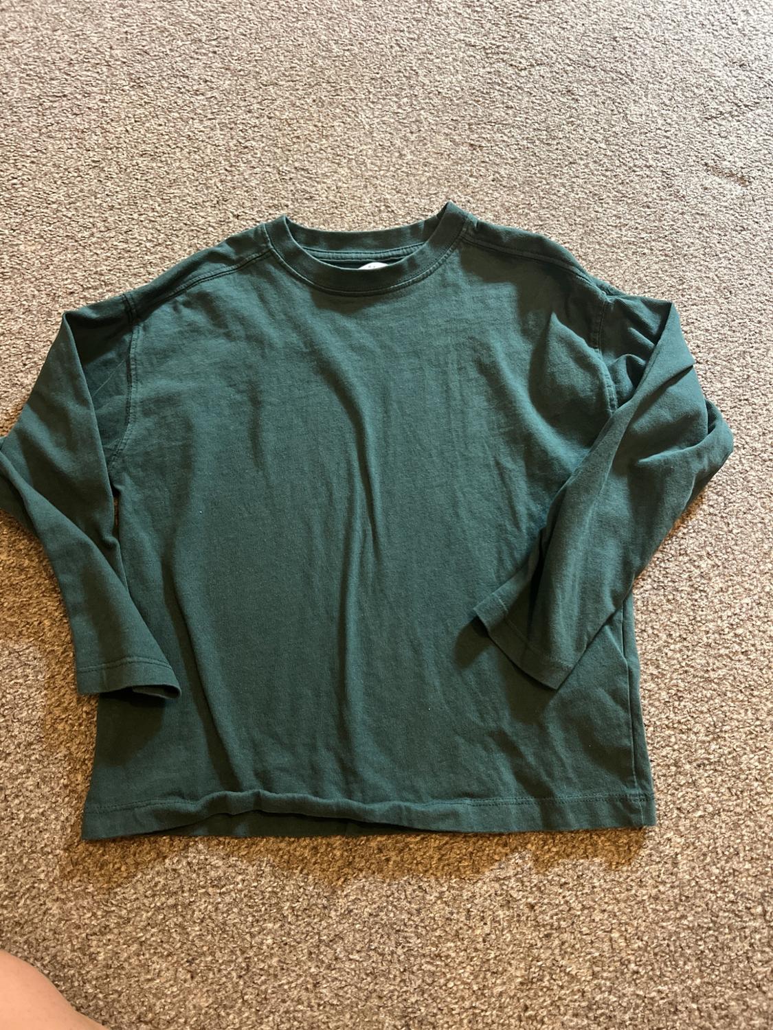 Dark green long-sleeved casual T-shirt with a round neck, laid out on a textured beige rug.