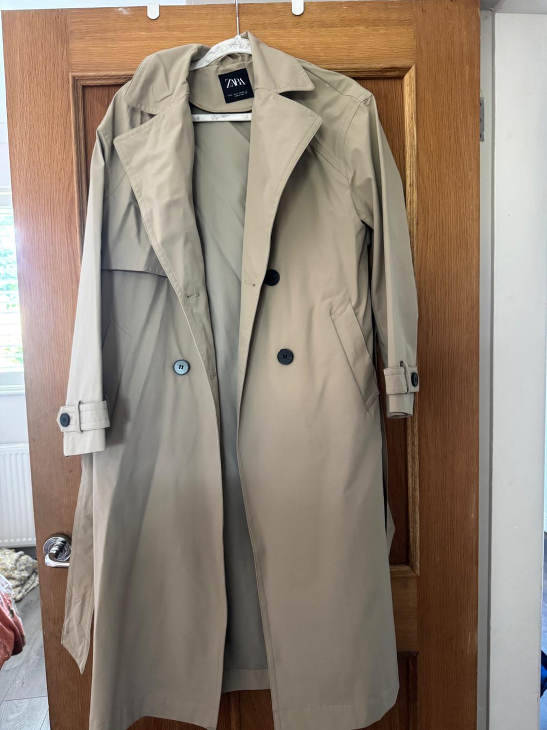 Beige trench coat hanging on a wooden door. It shows the ZARA label, dark buttons and cuff straps.