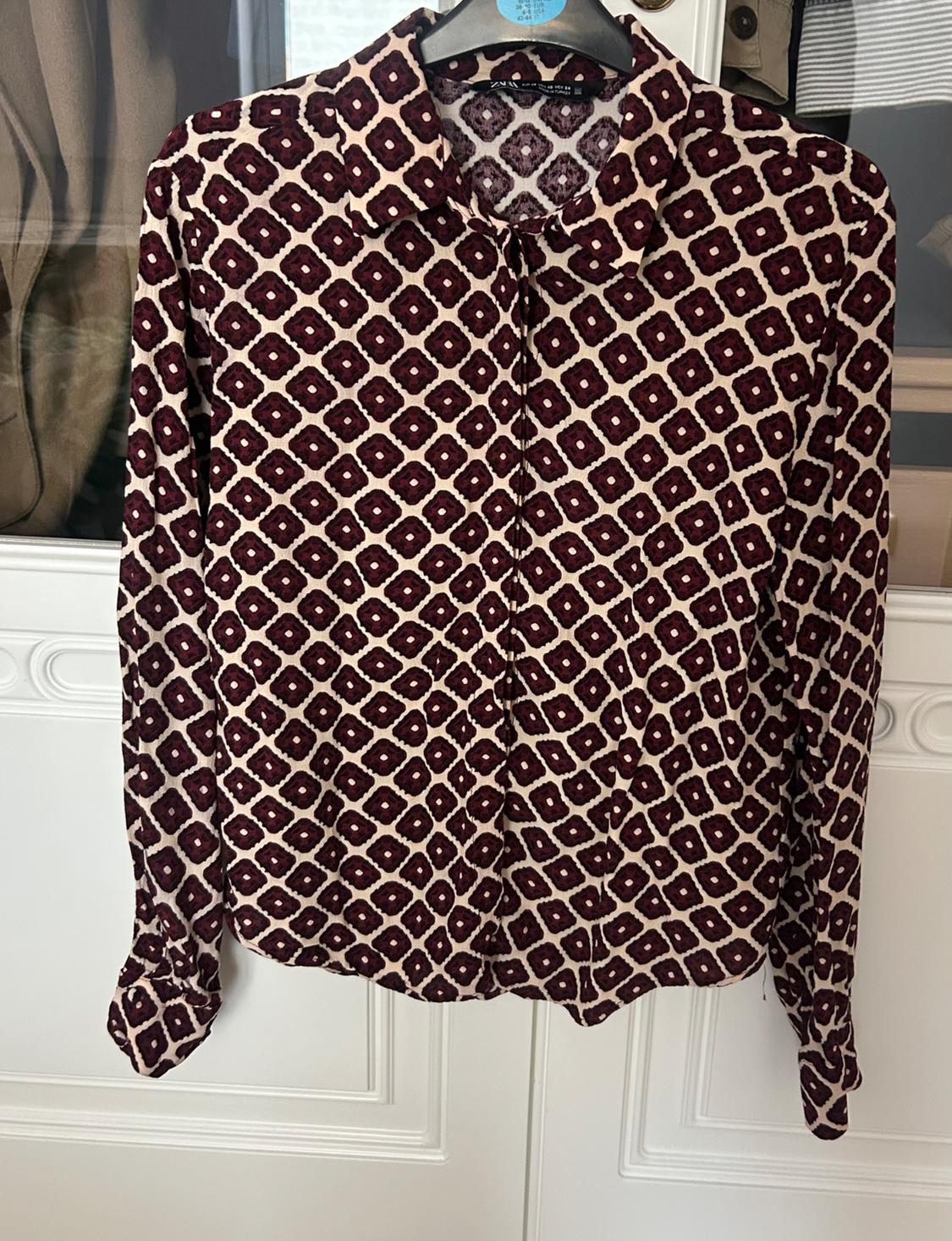 Long-sleeved shirt with a geometric print in burgundy and beige tones, hanging on a hanger in front of a white door.