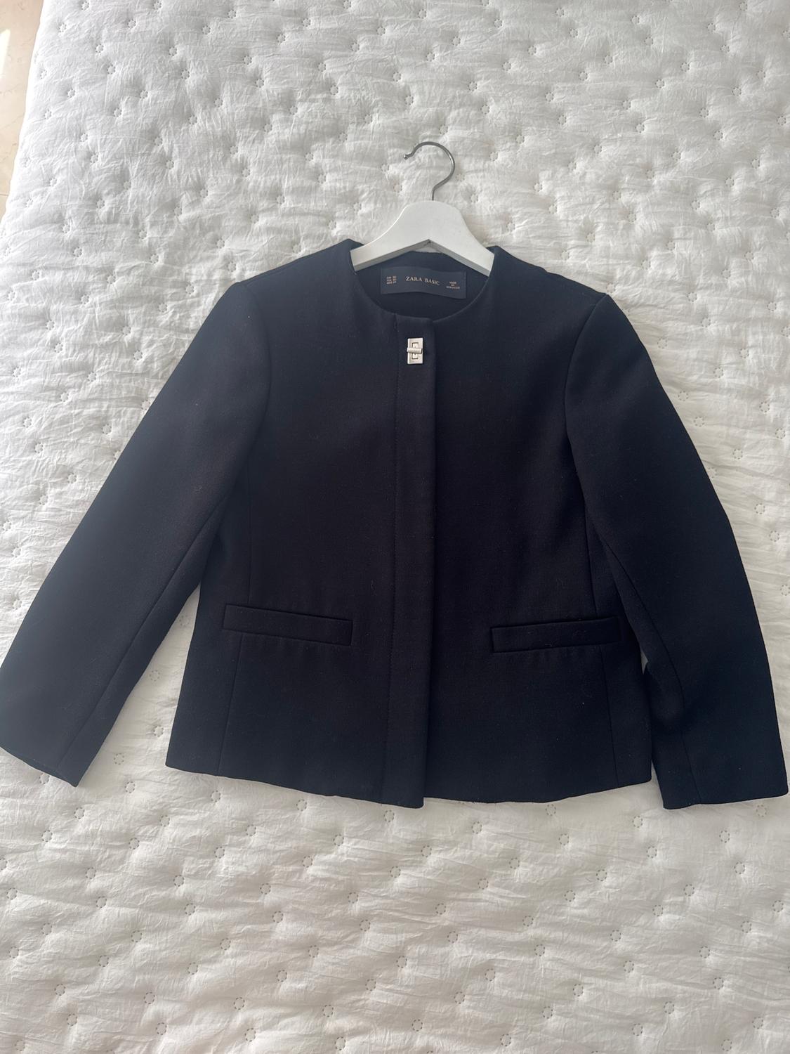 Black Zara Basic jacket hanging on a white hanger over a white quilted bedspread.